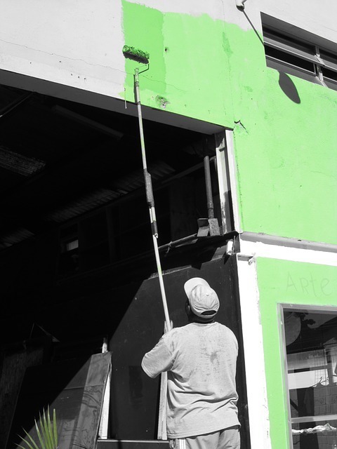 A person paints a building exterior with bright green paint using a roller on an extended pole, set against a grayscale background, conveying urban renewal.