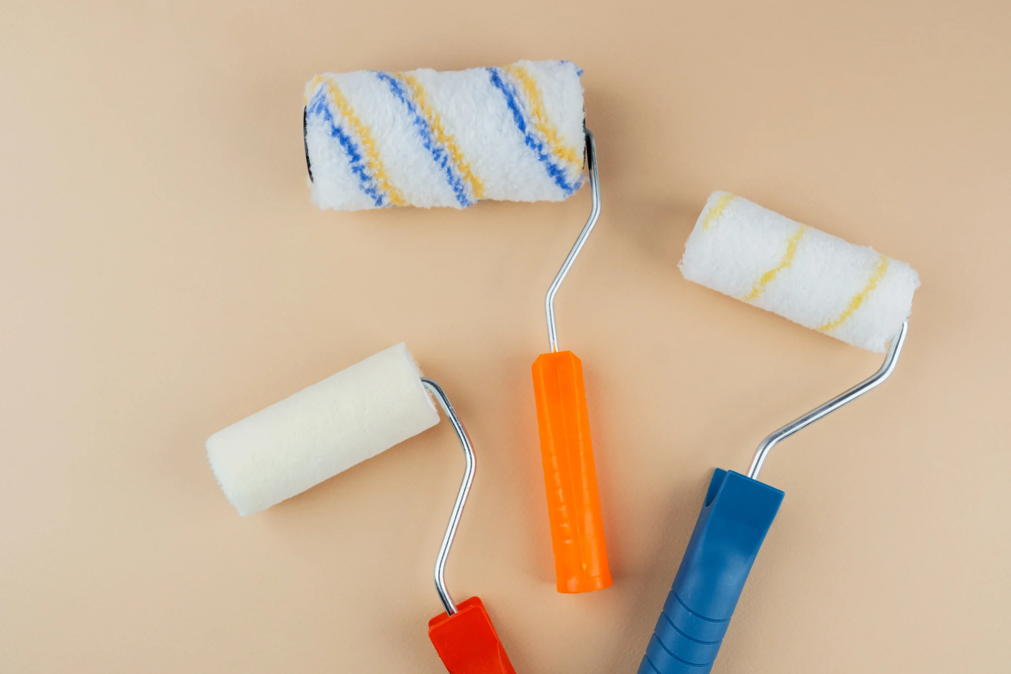Three paint rollers with colorful handles rest on a beige background. One roller is striped with blue and yellow, adding a vibrant touch.