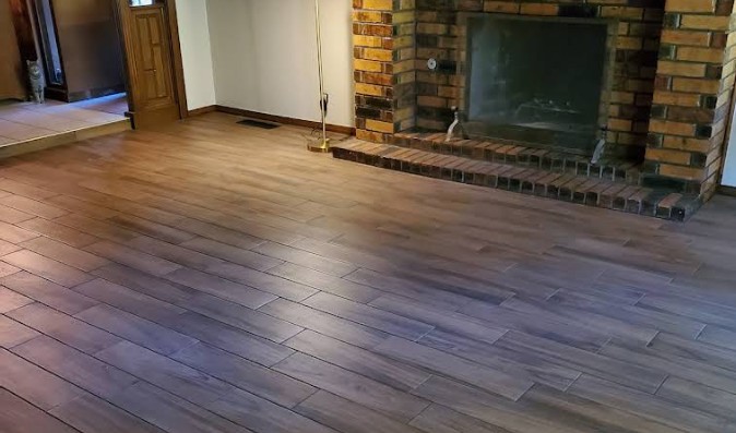 Spacious living room with wood-like tile flooring, brick fireplace, and a simple, cozy ambiance. A lamp stands in the corner, creating a warm tone.