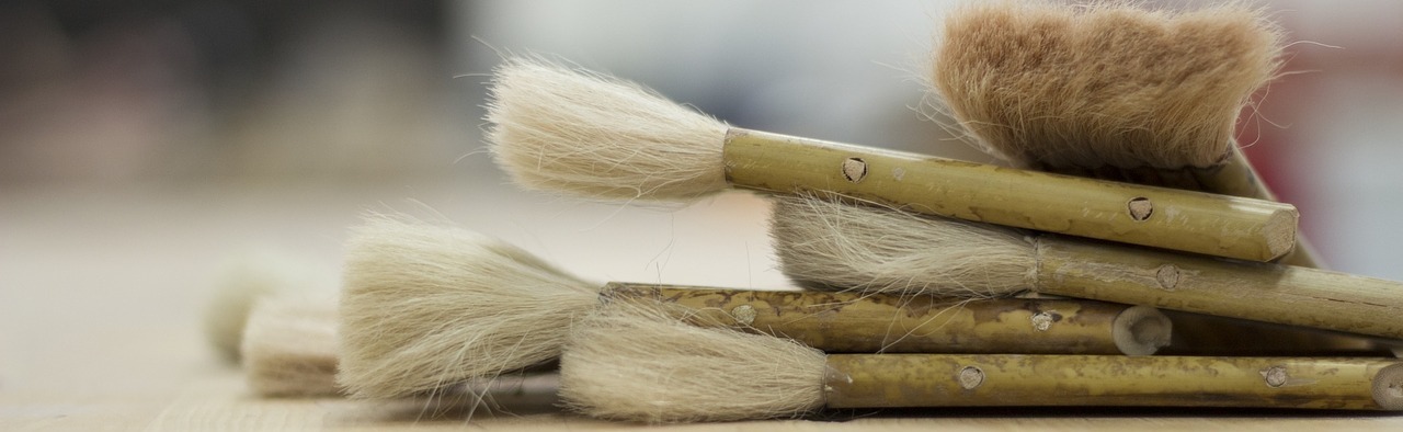 A stack of used paintbrushes with light-colored bristles and bamboo handles lies on a wooden surface, with a blurred background.